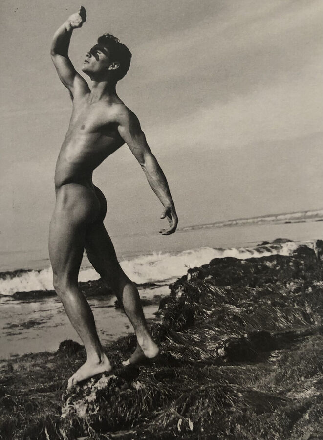 Black and white photograph of a naked muscular man seen from behind, standing on rocks by the sea with one arm raised to shield his eyes