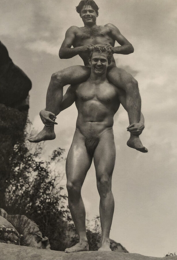 Black and white photograph of a naked muscular man standing on a rock outdoors, smiling, with a second man sitting on his shoulders