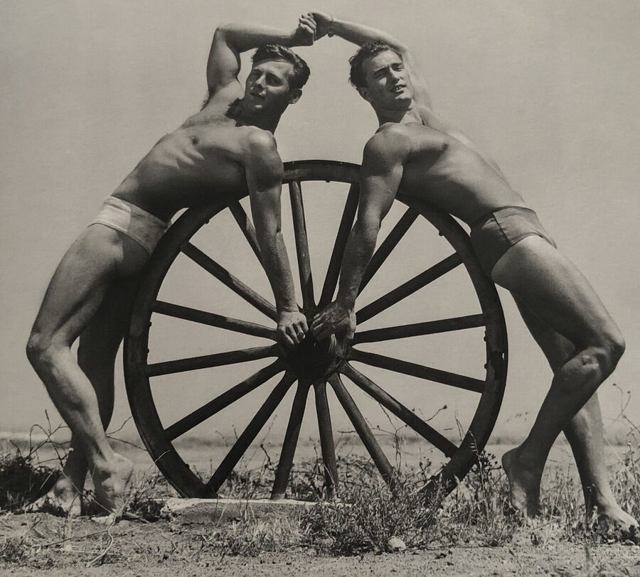 Two muscular men in white posing trunks leaning against a large wooden wagon wheel outdoors, their hands joined above their heads