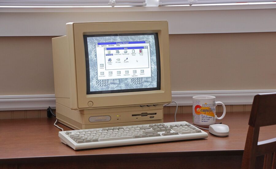 Beige 1990s desktop computer with a built-in monitor showing a Windows desktop, a keyboard, a mouse and a coffee mug on a wooden desk