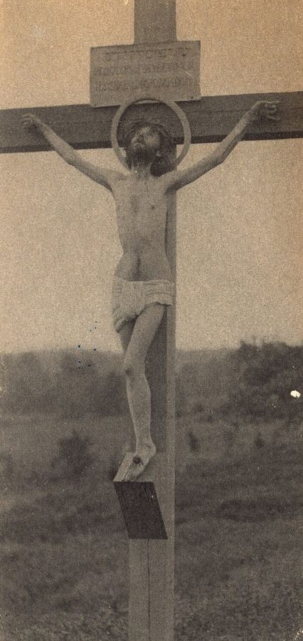 Photograph of a bearded man with a halo, naked except for a loincloth, hanging on a wooden cross in open countryside