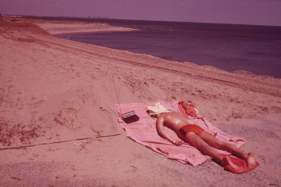 A man in red swimming trunks lies sunbathing on a pink towel on a sandy beach beside the sea
