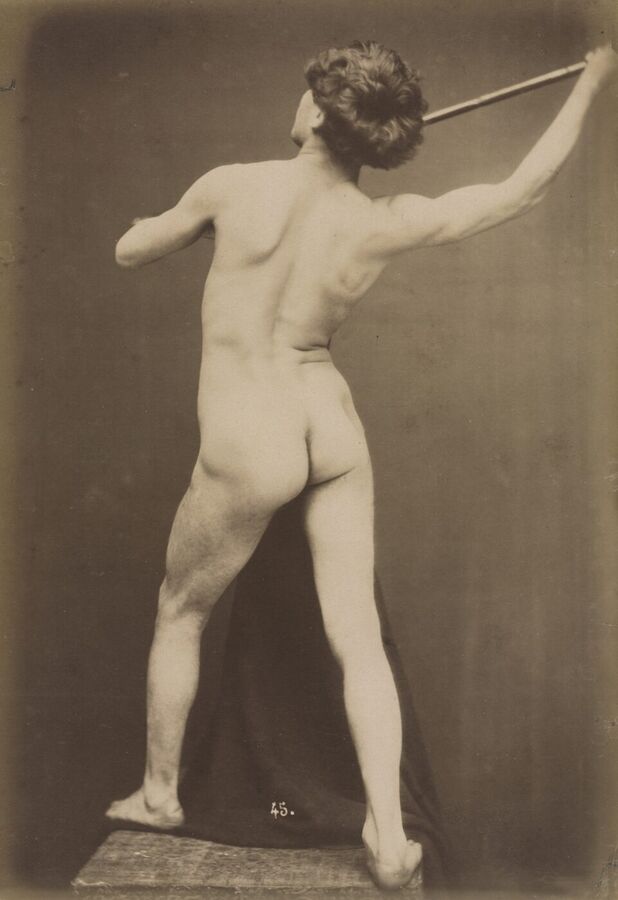 Photograph of a naked man seen from behind, standing on a wooden block and holding a long staff raised above his head