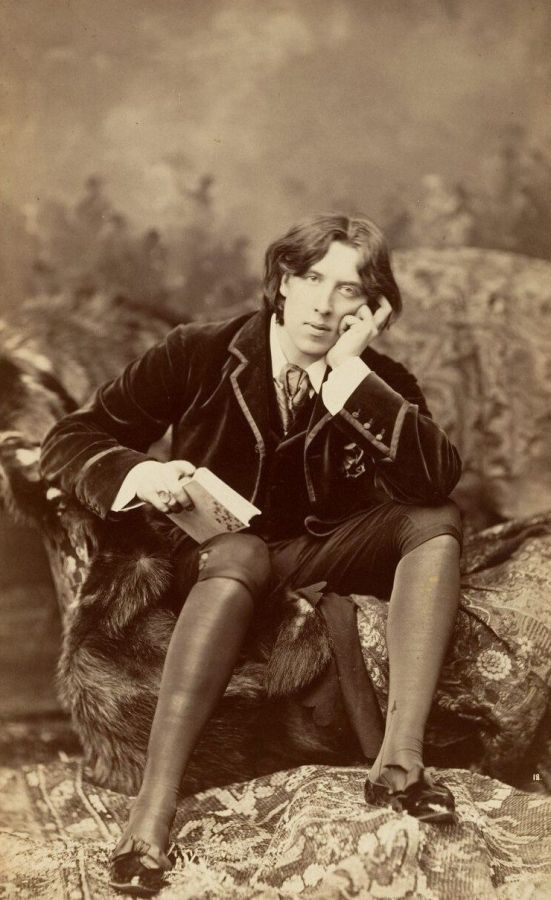 Photograph of Oscar Wilde seated on a fur-covered sofa, holding a book, in a velvet jacket and knee breeches