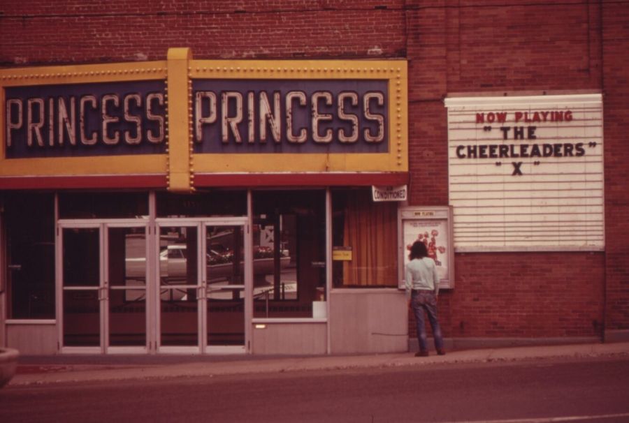 A man with long hair looks at a poster outside the Princess cinema, whose marquee board reads Now playing The Cheerleaders X