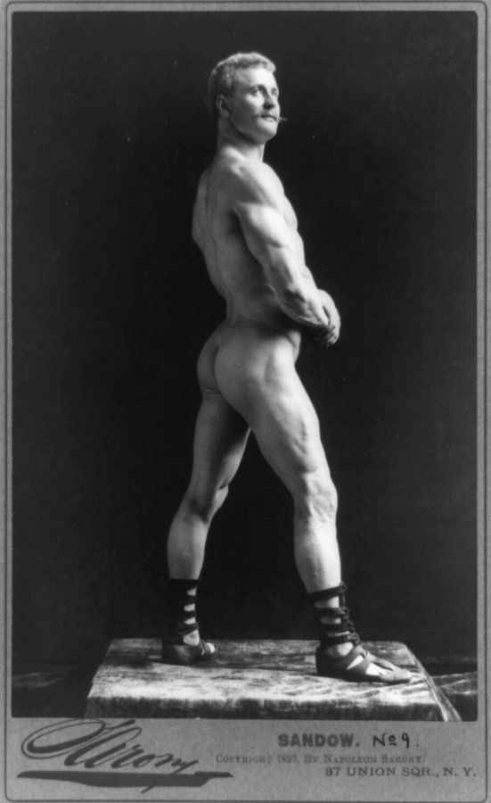 Photograph of Eugen Sandow standing naked on a plinth, seen in profile from behind with his hands clasped in front, wearing only laced sandals