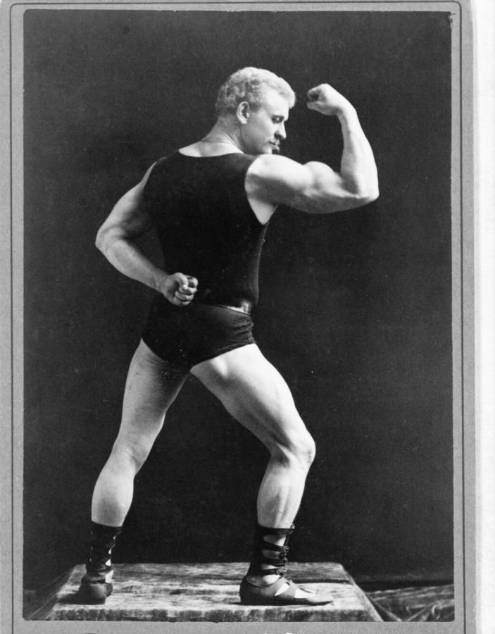 Photograph of the strongman Eugen Sandow in dark trunks and vest, flexing his right arm on a small plinth