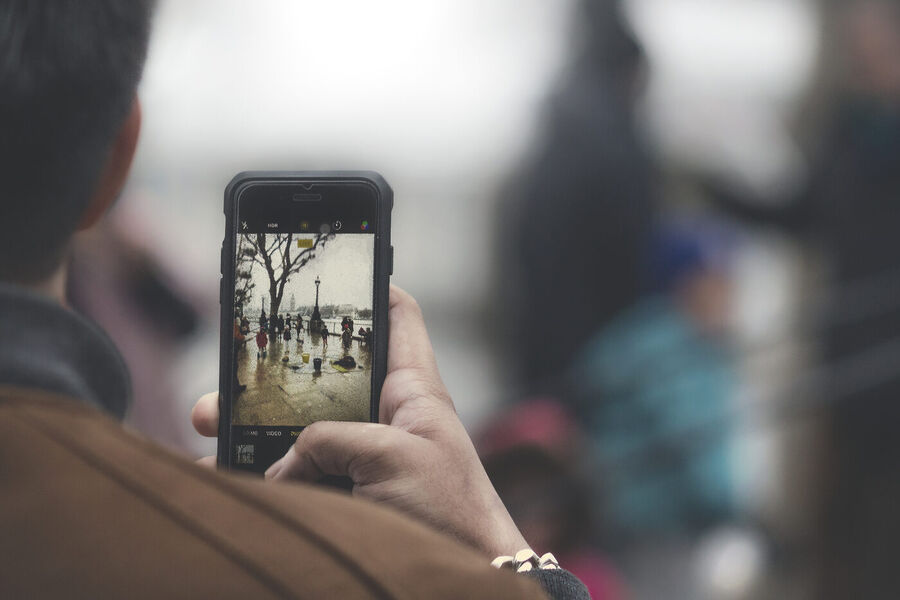 A hand holding a smartphone up to photograph a street scene, with the image visible on the phone's screen