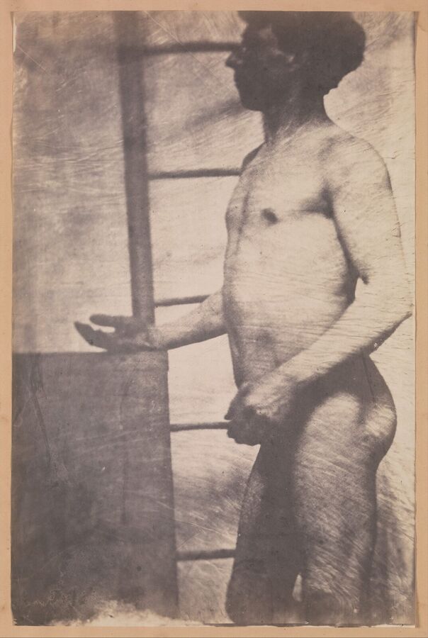 Faded brown photograph of a naked man standing in profile in front of a window frame, one hand held out