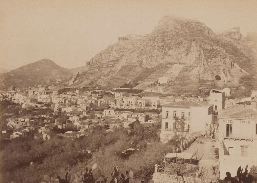 Old photograph of the town of Taormina on a hillside below a rocky mountain, with the Hotel Timeo in the foreground