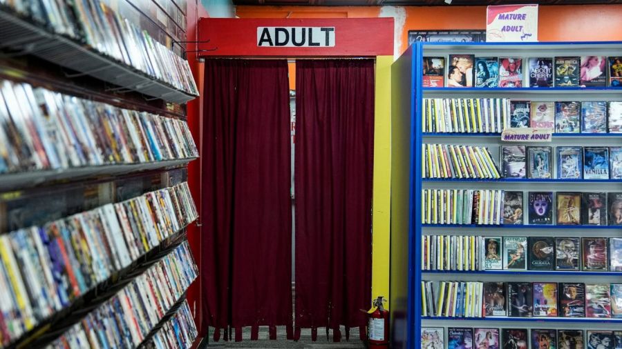 A doorway hung with dark red curtains under a sign reading ADULT, between shelves of DVDs in a video store