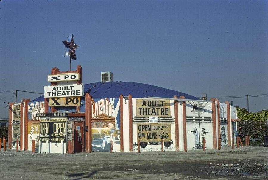A round, single-storey adult cinema painted with figures and signs reading Adult Theatre, XPO and Live Nude, under a clear blue sky