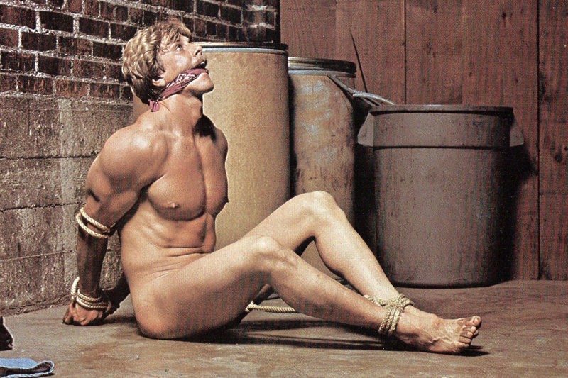 A naked muscular man with blond hair sits on a concrete floor against a brick wall, gagged with a red bandana, his wrists and ankles tied with rope
