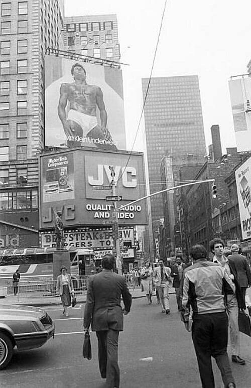 Black and white street photograph of Times Square with a giant billboard of a muscular man in white briefs above a JVC sign, and people crossing the street below