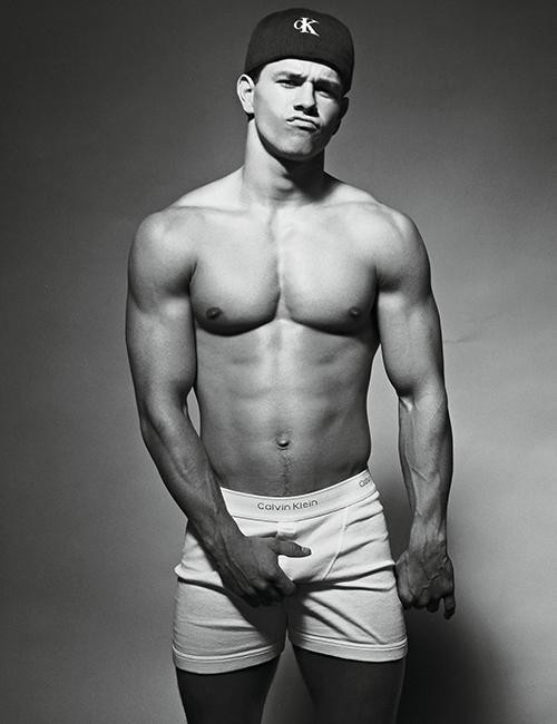 Black and white advertisement of a smiling muscular young man, bare-chested, in white Calvin Klein boxer briefs, with the Calvin Klein logo at the bottom