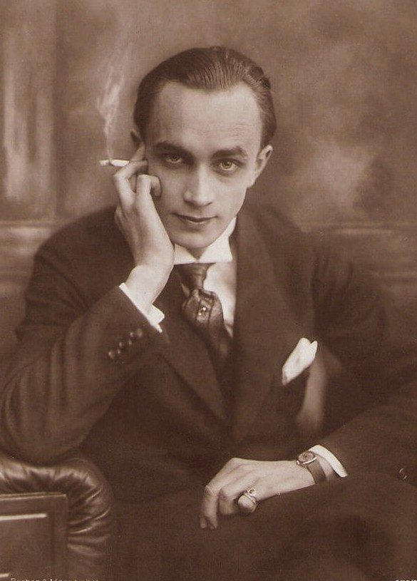 Sepia studio portrait of a young man in a dark suit and tie, sitting with his head resting on one hand, a cigarette between his fingers