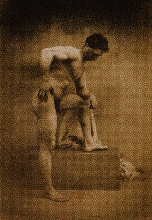 Sepia photograph of a muscular naked man leaning forward with one foot on a wooden block and his hands resting on his raised knee