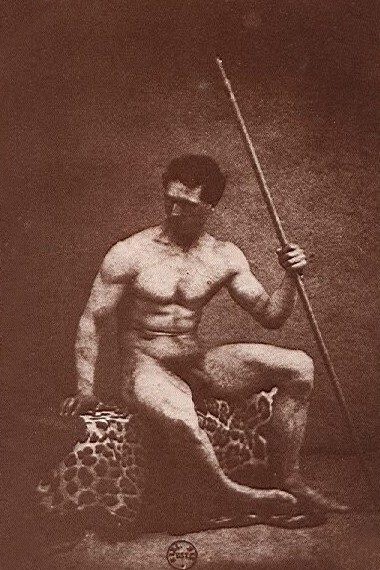 Brown-toned photograph of a muscular naked man seated on a leopard skin, holding a long staff upright in one hand