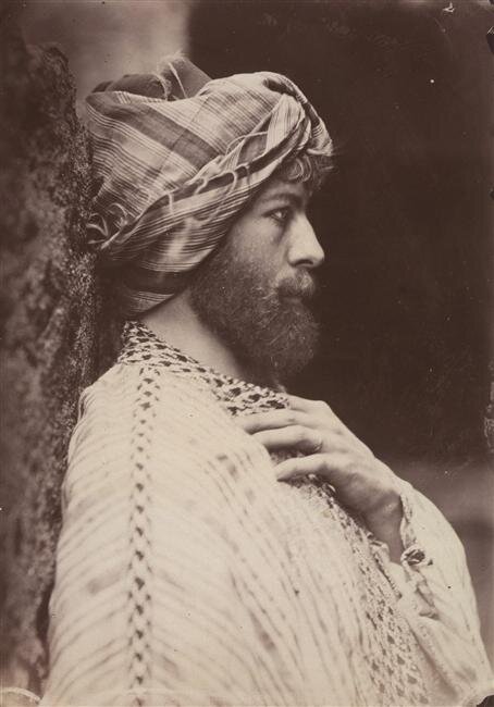 Sepia photograph of a bearded man in profile wearing a striped turban and a patterned robe, leaning against a rock