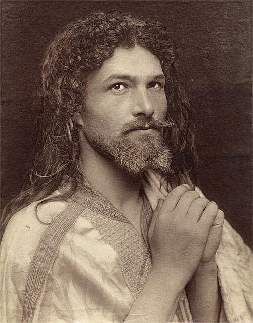 Sepia photograph of a man with long curly hair and a beard, hands joined in prayer, wearing a light embroidered robe