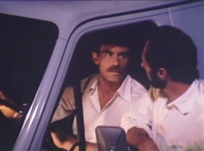At night, a moustached man in a white shirt sits at the wheel of a pale blue truck, talking through the window with a bearded man leaning in from the passenger side