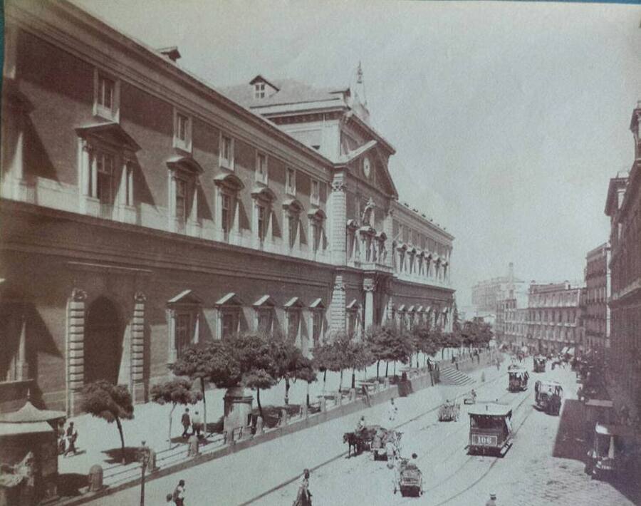 Long arcaded facade of the Naples museum along a wide street with trams, carriages and people walking