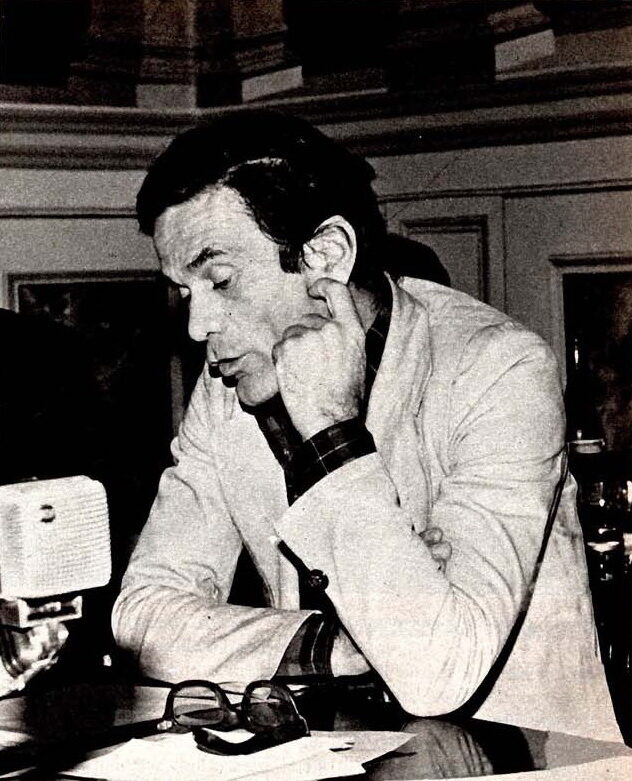 Black and white photograph of a dark-haired man in a light jacket leaning on a table, his fist against his cheek, with a microphone and a pair of glasses in front of him