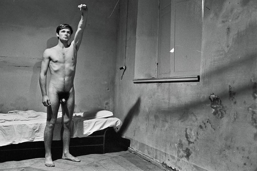 Black and white photograph of a naked young man standing by a narrow bed in a bare room with stained walls, his right arm raised above his head with the fist clenched