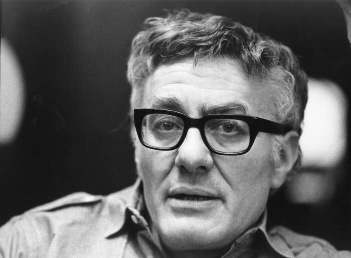 Black and white close-up of a man with greying wavy hair and thick black-rimmed glasses, speaking