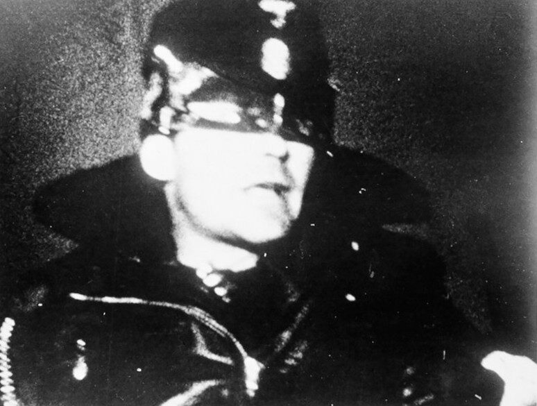 Black and white film still of a man in a shiny leather peaked cap and a black leather jacket with zips