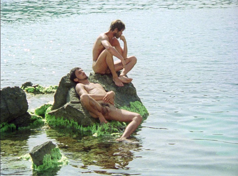 Two naked young men on a rock covered in green seaweed in shallow sea water, one sitting with his chin on his hand, the other lying back with his eyes closed