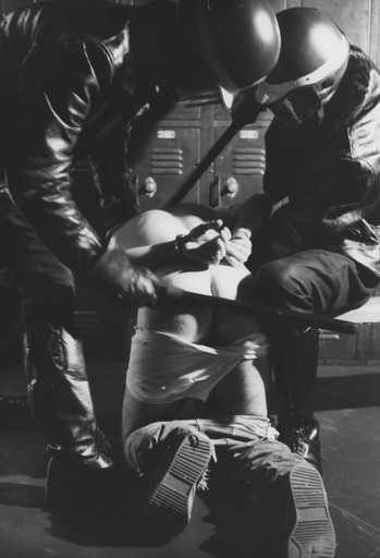 Black and white still of two men in motorcycle helmets and leather jackets in a locker room holding down a third man bent forward, his hands tied behind his back and his trousers pulled down