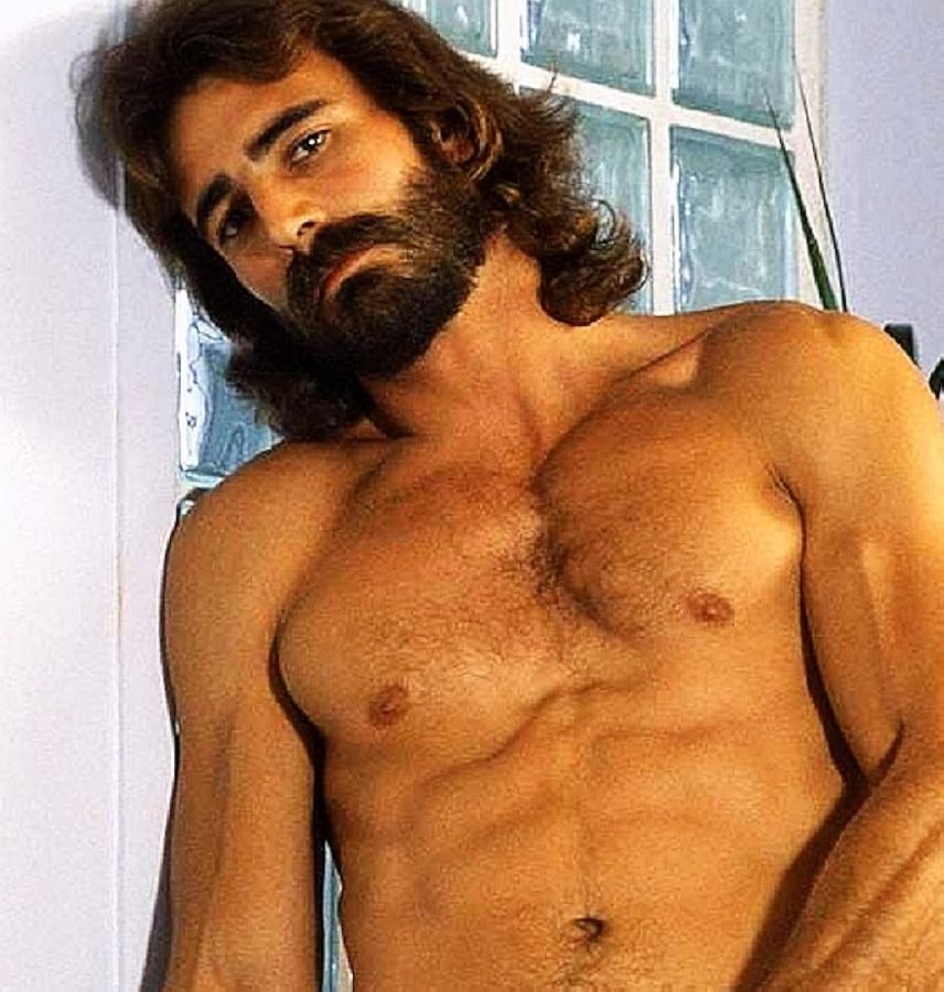 Al Parker, bare-chested, with long dark hair and a full beard, leaning back against a glass-brick wall