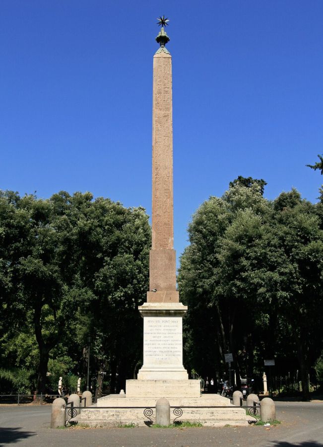 A tall red granite obelisk with hieroglyphs, topped by a metal star, standing on a stone pedestal in a park with trees and a blue sky