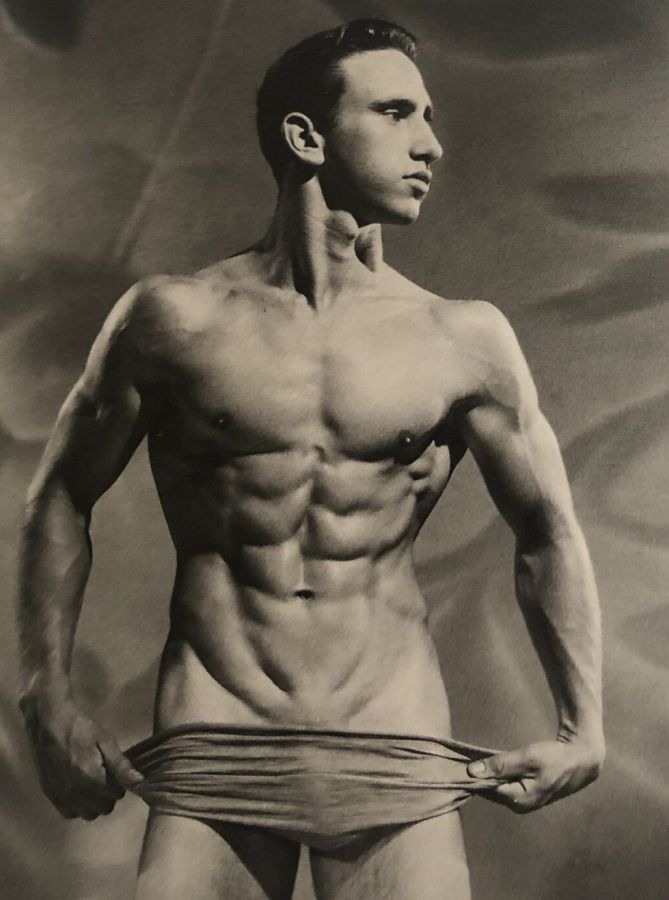 Black and white studio photograph of a lean muscular man with a defined abdomen pulling down the waistband of a cloth wrap below his navel