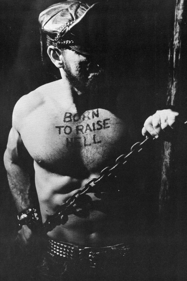 Black and white photograph of a bearded, shirtless man in a leather cap and studded belt holding a chain, with the words Born to Raise Hell written across his chest