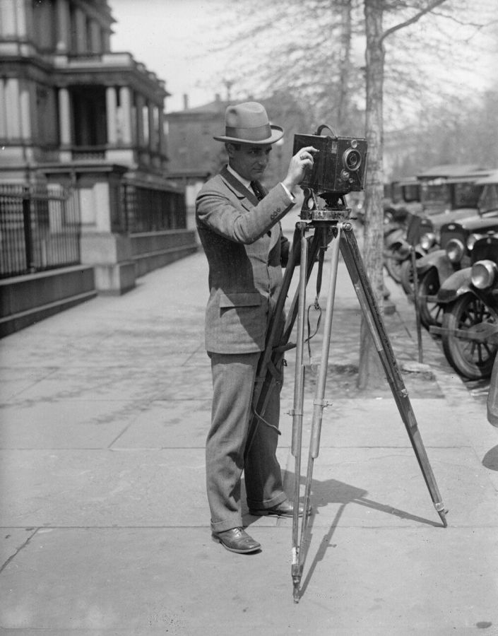 Black and white photograph of a man in a hat filming with a hand-cranked movie camera on a tripod on a city pavement, with parked cars behind him
