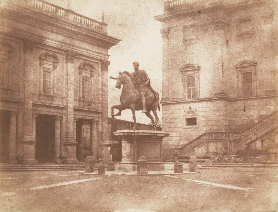 Old sepia photograph of a Roman square with a bronze statue of an emperor on horseback in the centre, between two classical palaces with balustrades