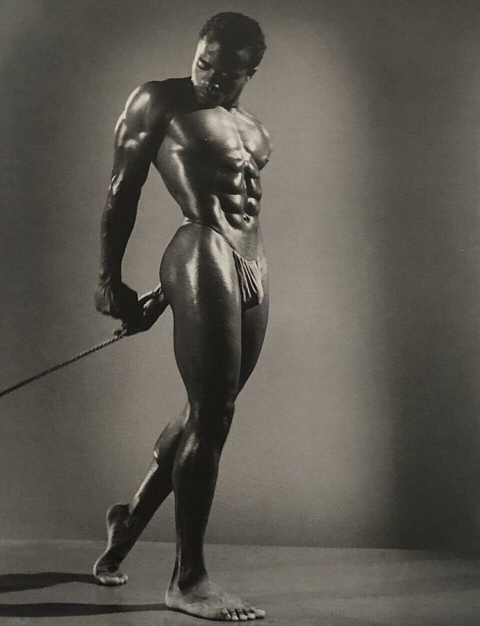 Black and white studio photograph of a muscular Black man in a small posing pouch, standing on one leg with his weight shifted and holding a long rod