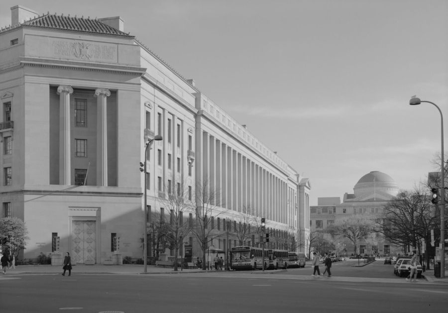 Black and white photograph of a long neoclassical government building with tall columns along a wide avenue, buses at the kerb and a domed building in the distance