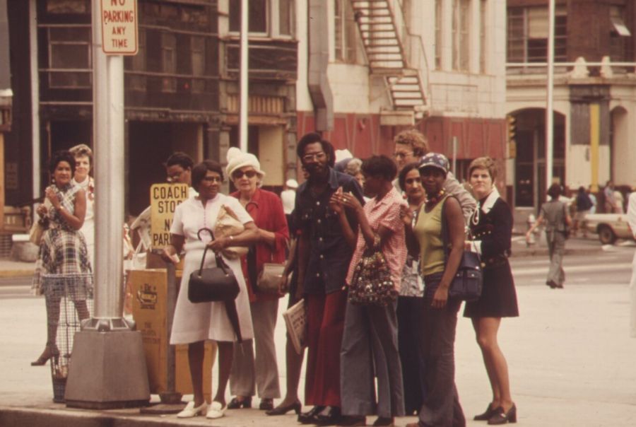 Men and women in 1970s clothes waiting at a bus stop on a street corner in downtown Atlanta, with older brick buildings behind them
