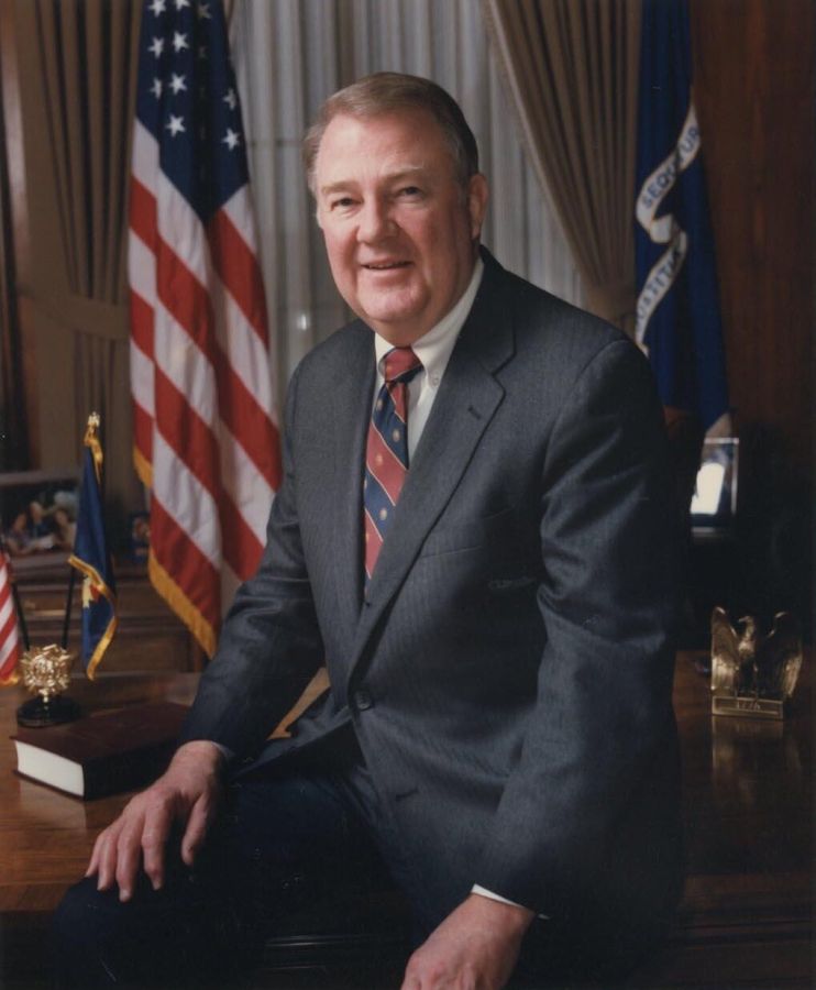 Colour portrait of a heavy-set grey-haired man in a dark suit and striped tie, seated on the edge of a desk in front of American flags