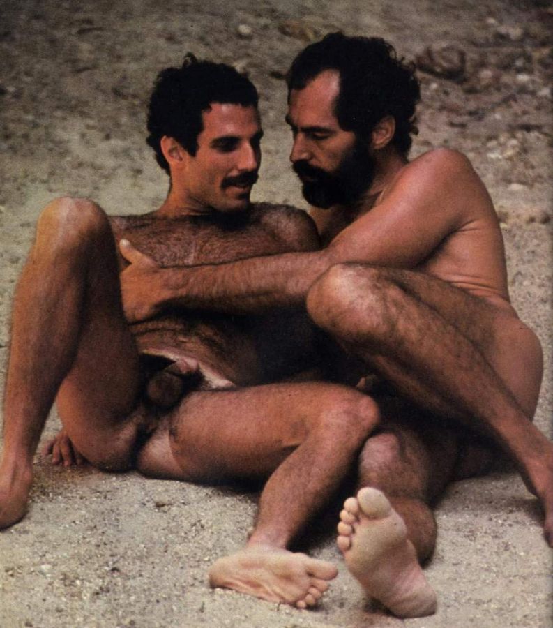 Two naked, hairy, moustached men sit entwined on the sand, one holding the other from behind