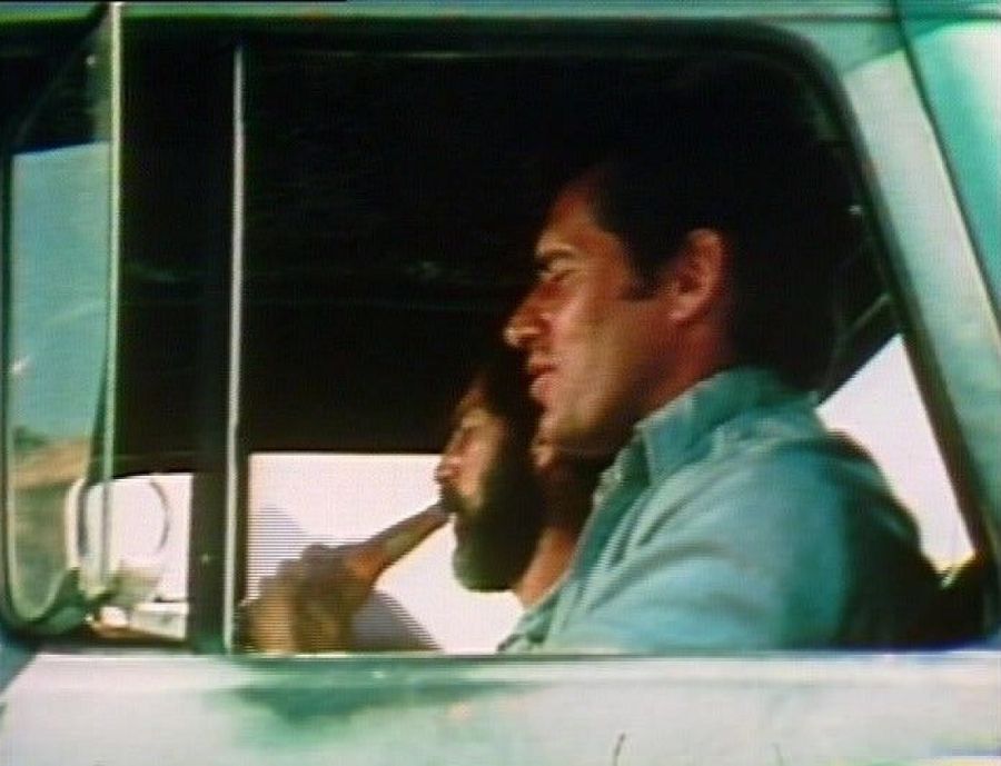 Two men in the cab of a pale green truck on the road, a clean-shaven driver in a blue shirt and a bearded passenger drinking from a bottle