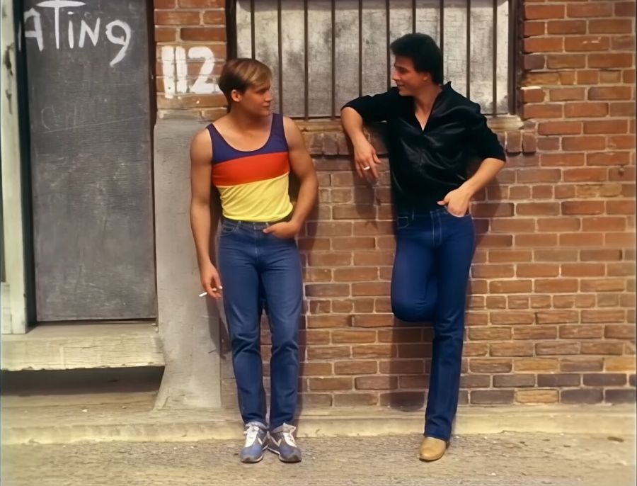 Two clean-cut young men in jeans lean against a brick wall in the street, one in a striped tank top, the other in a dark shirt with a cigarette