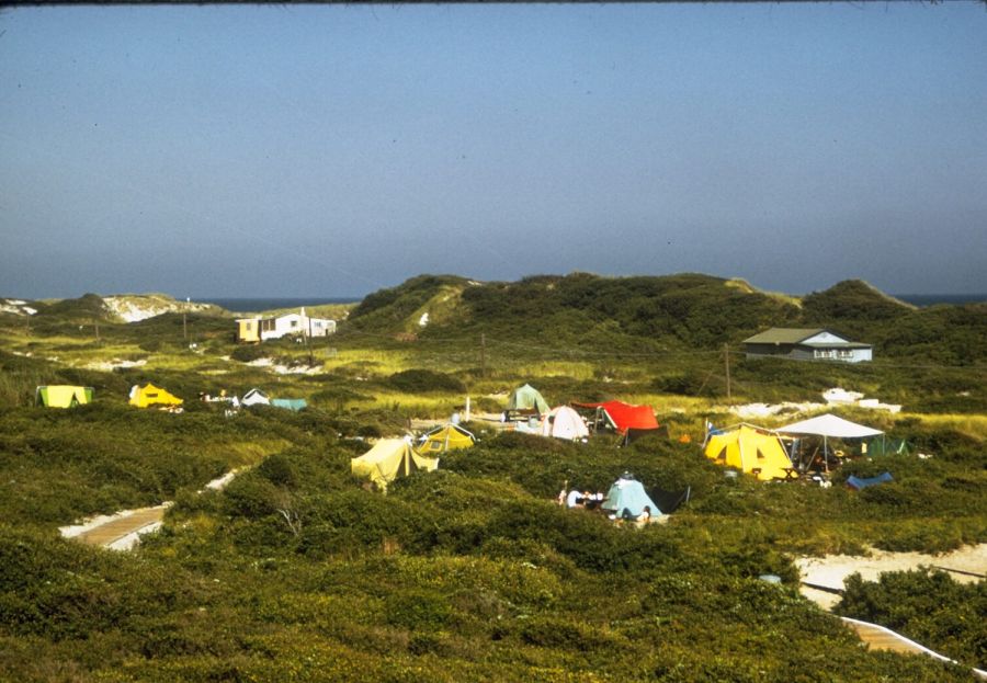 Small tents and a few low buildings scattered among green, scrub-covered dunes, with the sea on the horizon under a clear sky