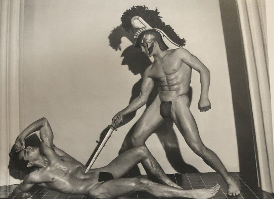 Black and white studio photograph of a muscular man in a crested helmet and posing strap holding a sword over a second man lying on the floor