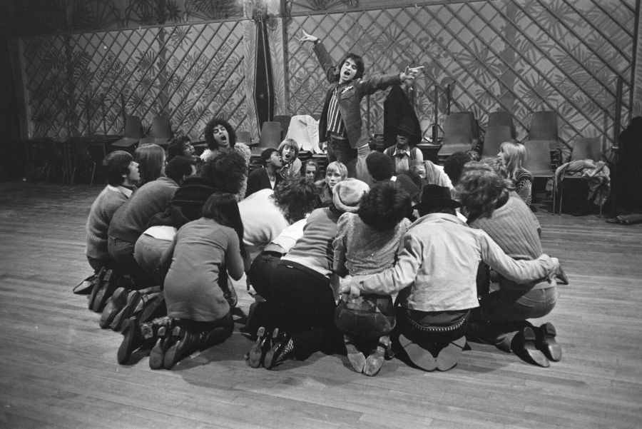Black and white photograph of a young cast in casual clothes crowded together on the floor of a rehearsal room, with one actor raised above the group, arms outstretched