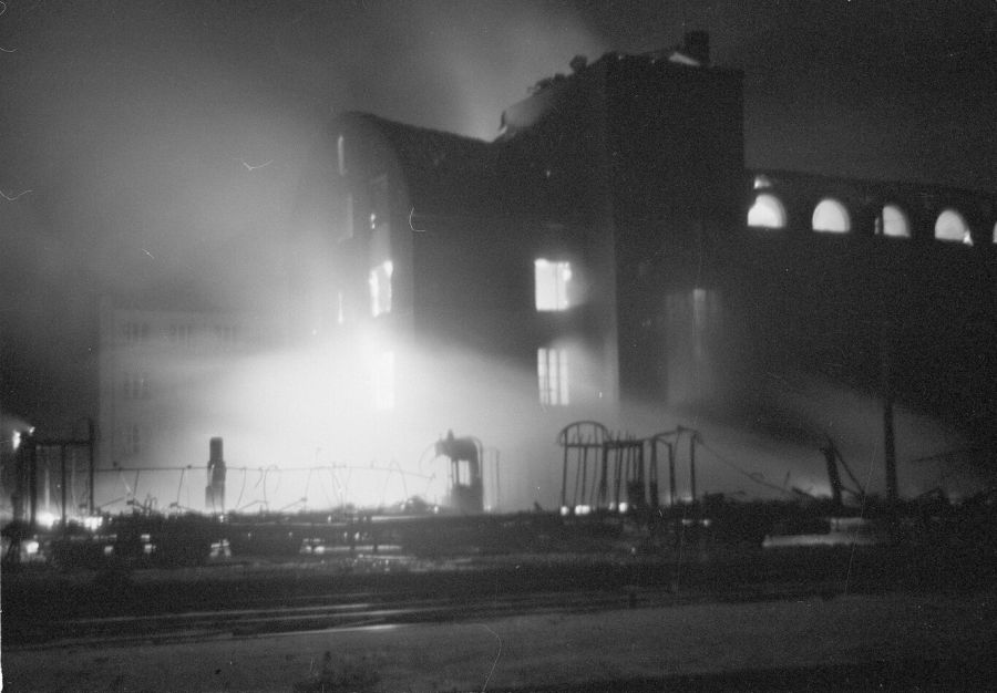 Night photograph of a large stone building with arched windows, lit by fire and wrapped in smoke, with railway tracks in the foreground