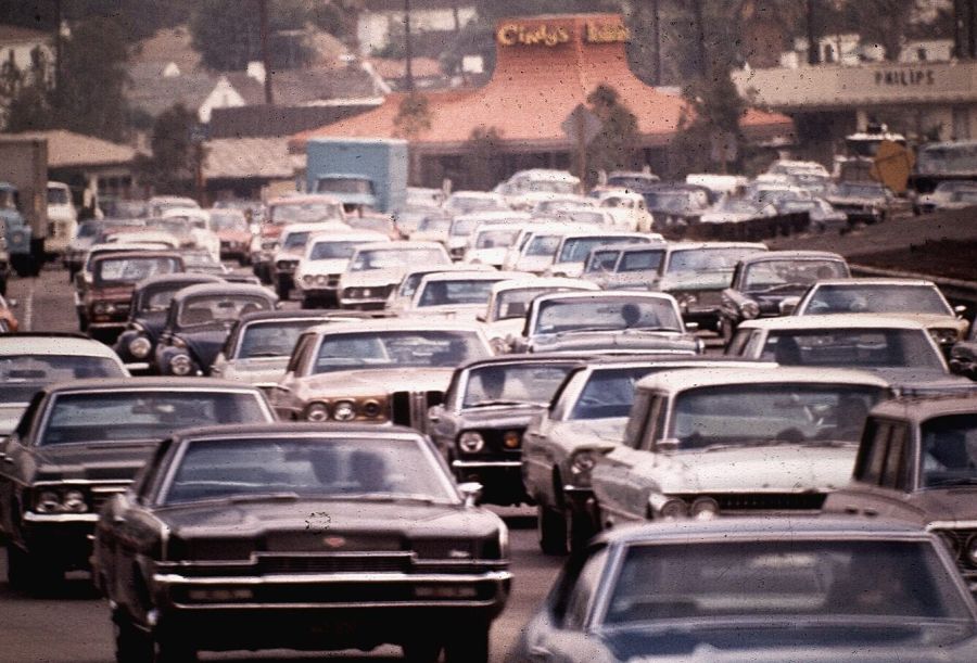 A jam of large American cars filling every lane of a freeway, with a restaurant with a tall orange roof and low buildings on the hillside behind